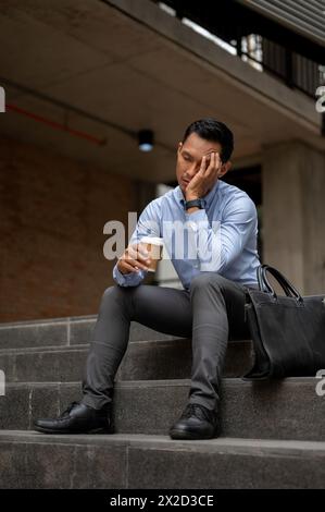 Un homme d'affaires millénaire asiatique stressé, paraissant fatigué et malheureux, est assis sur les marches à l'extérieur d'un bâtiment, tenant une tasse de café et couvrant son visage Banque D'Images