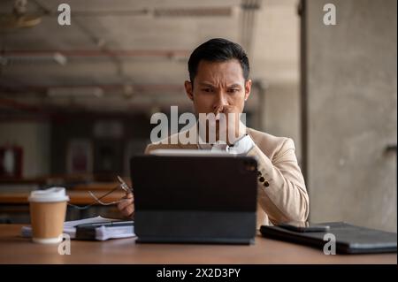 Un homme d'affaires millénaire asiatique stressé et réfléchi regarde son écran de tablette numérique avec une expression sérieuse, confronté à un problème pendant le travail, s. Banque D'Images