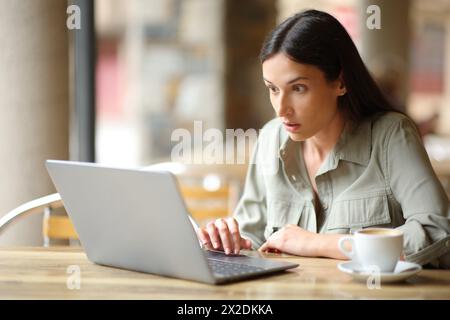 Femme stupéfaite regardant les nouvelles sur un ordinateur portable dans une terrasse de bar Banque D'Images