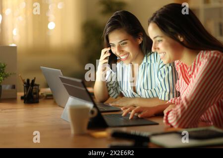 Télétravailleurs heureux utilisant plusieurs appareils et parlant au téléphone dans la nuit à la maison Banque D'Images