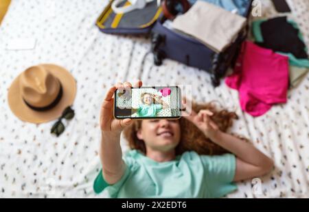 Femme allongée sur le lit à l'hôtel et prend un selfie. Messagerie photo avec des amis en voyage. Banque D'Images