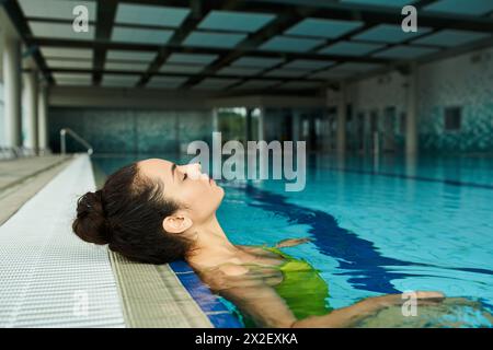 Une jeune et belle femme brune en maillot de bain flotte sans effort dans une piscine intérieure spa. Banque D'Images