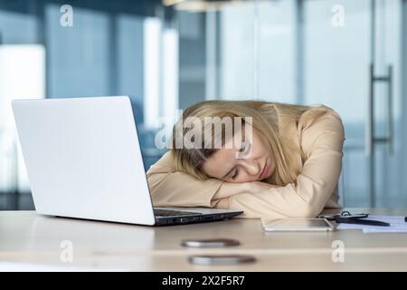 Jeune femme blonde prise dans un moment d’épuisement, dormant à son bureau entourée d’objets de travail, dont un smartphone, des stylos et un ordinateur portable. Banque D'Images