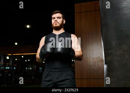 Un bel homme barbu portant des gants de boxe se tient confiant à côté d'un sac de frappe dans une salle de gym. Banque D'Images