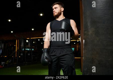 Un homme barbu dans un débardeur noir jette des coups de poing sur un sac de punching suspendu dans un cadre de gymnastique. Banque D'Images