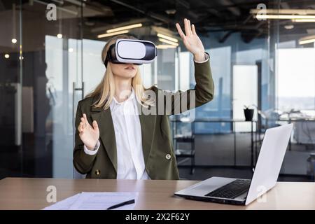 Une femme d'affaires professionnelle utilisant un casque de réalité virtuelle à son bureau dans un environnement de bureau contemporain, démontrant l'intégration de la technologie de pointe dans le lieu de travail. Banque D'Images