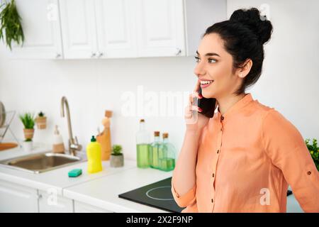 Une femme élégante en tenue décontractée se tient dans une cuisine, discutant sur un téléphone portable tout en travaillant. Banque D'Images