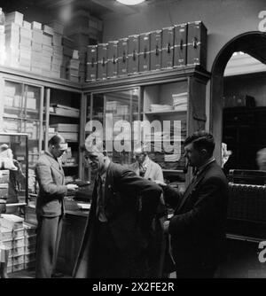 LONDON MERCHANT SEAMEN'S HOME AND RED ENSIGN CLUB, 1942 - le marin marchand George Lappin tente un manteau, aidé par son collègue Albert John Burrett, dans la boutique bien équipée du Merchant Seamen's Home and Red Ensign Club. Le magasin fournit des vêtements et du matériel, qui peuvent être vus empilés sur des étagères autour de la pièce. Le Storeman et un autre marin peuvent également être vus en arrière-plan Banque D'Images