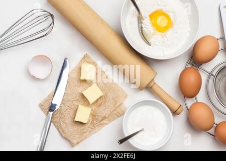 Faire dorer les œufs sur un support en métal. Beurre et couteau sur papier. Jaune d'œuf avec farine et cuillère dans un bol. Lait dans le bol et rouleau à pâtisserie sur la table. Fond blanc. Banque D'Images