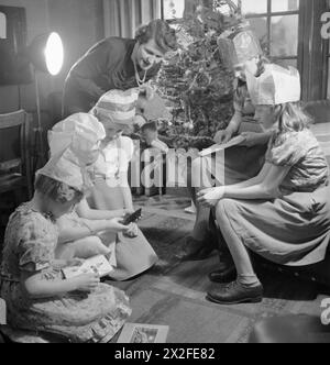 FÊTE DE NOËL POUR LA FILLE du SOLDAT DEVEREUX : NOËL EN TEMPS DE GUERRE, PINNER, MIDDLESEX, DÉCEMBRE 1944 - Mme Devereux regarde sa fille Jean (troisième à partir de la gauche) et ses amis ouvrir leurs cadeaux de Noël, vers la fin de la fête de thé de Noël qu'ils ont à leur domicile à Pinner. Beaucoup de filles portent des chapeaux de fête en papier. Le sapin de Noël à l'arrière-plan est un cadeau du mari de Mme Devereux, qui sert actuellement dans l'armée outre-mer. L'arbre a été acheté par le biais de la « Gifts to Home League » du YMCA. Mme Devereux tient un ours en peluche Banque D'Images