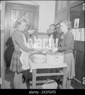 LES JARDINS DU VILLAGE NOURRISSENT LES ÉCOLIERS : PRODUCTION ALIMENTAIRE À KNIGHTON-ON-TEME, WORCESTERSHIRE, ANGLETERRE, Royaume-Uni, 1943 - dans le coin d'une salle de classe utilisée comme salle à manger à l'école du village de Knighton-on-Teme, trois enseignants préparent le repas du midi. Deux des filles plus âgées ramassent les assiettes et portent les repas aux tables. Les légumes consommés aujourd'hui ont été vendus à l'école par des membres de l'Association locale des produits du village Banque D'Images