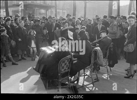 PARIS, PRINTEMPS 1945 : LA VIE QUOTIDIENNE À PARIS LIBÉRÉ, FRANCE, 1945 - Un grand groupe de personnes, y compris des enfants et des soldats américains et britanniques, se sont rassemblés autour d'un trio de musiciens pour entendre une nouvelle chanson sur la place de la Madelaine, Paris, France. Le soleil brille sur les musiciens (un accordéon, une batteur et un violoniste) alors qu'ils sont dirigés par une femme plus âgée, qui se prépare à chanter. Un petit kiosque à journaux peut être vu en arrière-plan Banque D'Images