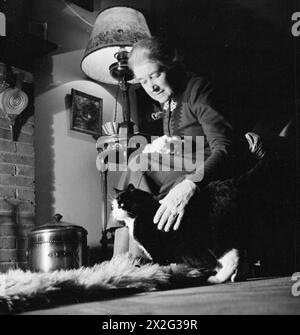 MME BUGLER VA À LA GUERRE : LA VIE QUOTIDIENNE À EAST DEAN, SUSSEX, ANGLETERRE, 1943 - à la fin d'une longue journée, Mme Bugler et son chat Tommy se détendent devant le feu chez elle à East Dean Banque D'Images