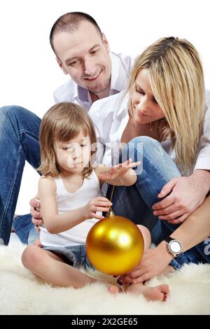 Père, mère et petite fille qui tient une grande boule de noël dorée assis sur le tapis blanc velu. Banque D'Images