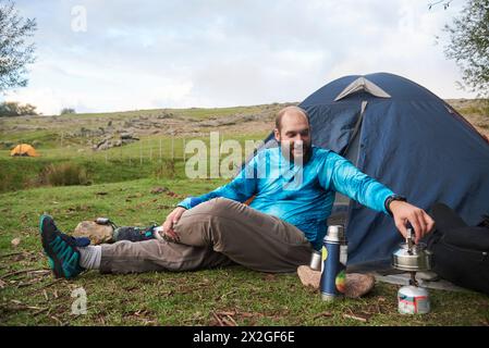 Jeune homme assis détendu dans un camp, devant une tente, souriant, chauffant l'eau sur son poêle. Concept : profiter de vacances en plein air, en contact Banque D'Images