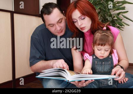 mère, père et petite fille en denim combinaison lit livre, plante d'intérieur, focus sur la fille Banque D'Images