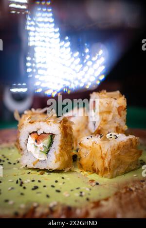 Un gros plan de rouleaux de sushi avec des graines de sésame sur une assiette verte éclairée par des lumières arc-en-ciel, prêt à être mangé avec des baguettes reposant sur l'assiette. Banque D'Images