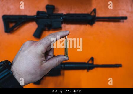 Armes de tir dans un champ de tir. Une cartouche de 5,56 x 45 mm dans la main d'un homme et un fusil m4a1 sur un mur orange. Banque D'Images