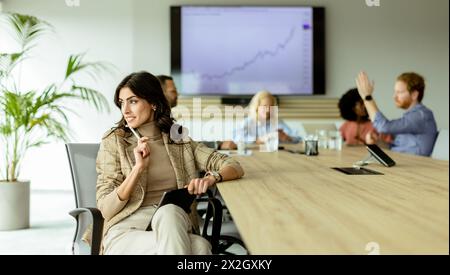 Femme d'affaires concentrée avec un bloc-notes réfléchit lors d'une réunion d'équipe, avec des collègues engagés dans la discussion en arrière-plan Banque D'Images