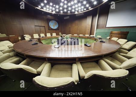 Auditorium avec table ronde en chêne, fauteuils beiges autour, écran blanc sur le mur. Banque D'Images