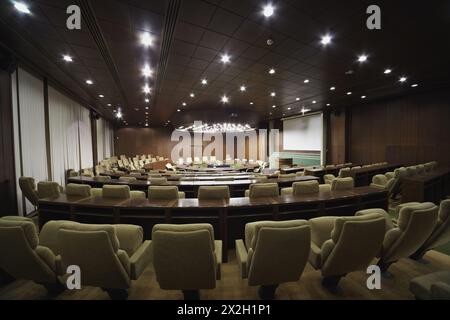 Salle de réunion avec table ronde en chêne, fauteuils beiges et rangées de bureaux et fauteuils. Banque D'Images