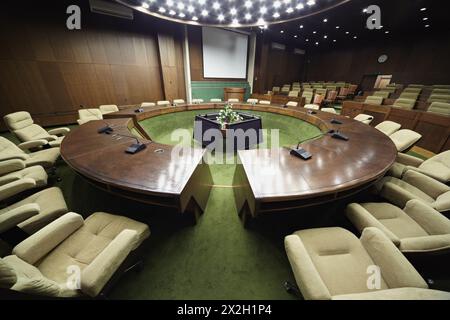 Auditorium avec table ronde en chêne et fauteuils beiges autour, rangées de fauteuils pour les participants à une réunion. Banque D'Images