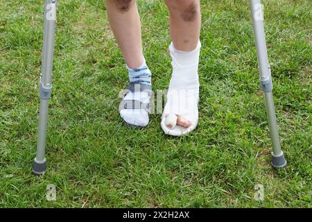 Jambes du petit garçon sur des béquilles ; jambe gauche en plâtre ; garçon debout sur l'herbe verte Banque D'Images