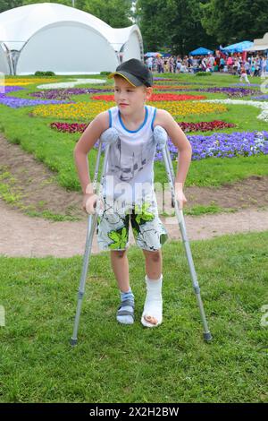 Triste petit garçon sur des béquilles dans le beau parc d'été ; jambe en plâtre Banque D'Images