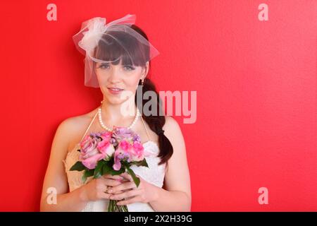 mariée portant une robe blanche et un voile court tient un bouquet de roses et regarde la caméra sur fond rouge Banque D'Images