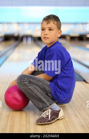 Garçon vêtu d'un T-shirt bleu avec boule rose se trouve sur le sol dans le club de bowling ; faible profondeur de champ ; corps entier Banque D'Images