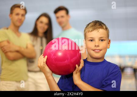 Garçon souriant vêtu d'un T-shirt bleu tient la balle rose dans le club de bowling ; deux hommes et une femme se tiennent derrière lui ; se concentrer sur le garçon ; faible profondeur de champ Banque D'Images