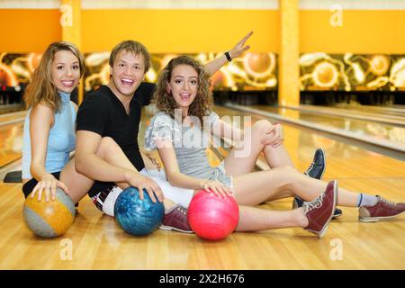 Deux belles filles heureuses et l'homme assis sur le sol avec des balles dans le club de bowling Banque D'Images