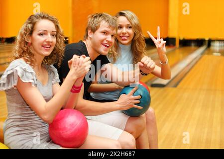 Jeune homme et deux belles filles heureuses s'assoient, tiennent des balles et félicitent quelqu'un dans le club de bowling ; concentrez-vous sur l'homme Banque D'Images