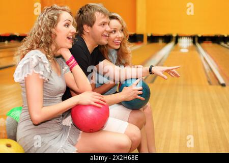 Jeune homme et deux belles filles heureuses s'asseyent, tiennent des balles et se demandent quelque chose dans le club de bowling ; concentrez-vous sur l'homme Banque D'Images