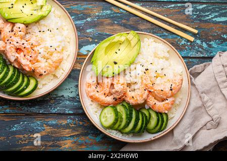 Deux bols en céramique blanche avec riz, crevettes, avocat, légumes et graines de sésame et baguettes sur fond de bois rustique coloré vue de dessus Banque D'Images