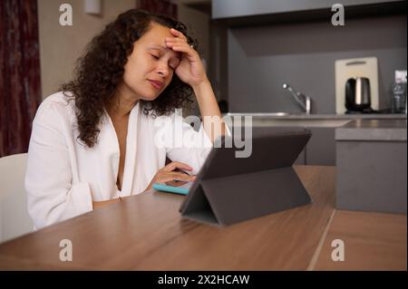 Femme d'affaires fatiguée en peignoir blanc à la maison, travaillant en ligne sur tablette, lisant les e-mails avec de mauvaises nouvelles, souffrant d'épuisement professionnel, assis à la table de la cuisine Banque D'Images
