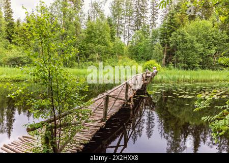 Vieille passerelle en bois au-dessus d'une rivière construite de bâtons d'arbre avec un homme assis sur un banc dans une forêt verdoyante en été, Mullsjoe, Suède Banque D'Images