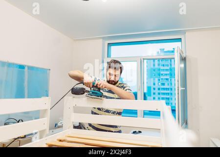 l'homme frotte le lit d'enfant en bois avec une ponceuse électrique ou un broyeur en enlevant la vieille peinture Banque D'Images