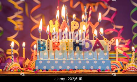 Bougies allumées de différentes couleurs en forme de lettres formant l'expression JOYEUX ANNIVERSAIRE, sur un fond flou violet avec des streamers Banque D'Images