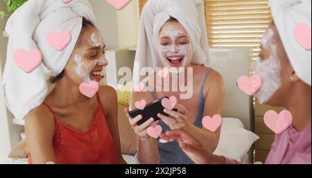 Image de coeurs roses flottant sur des amis féminins divers heureux appliquant des masques faciaux de beauté Banque D'Images