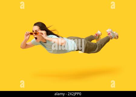 Jeune femme surprise dans des lunettes de soleil volant sur fond jaune Banque D'Images