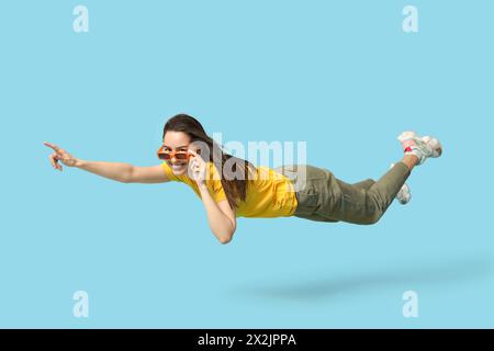 Surprise jeune femme dans des lunettes de soleil volant et pointant vers quelque chose sur fond bleu Banque D'Images