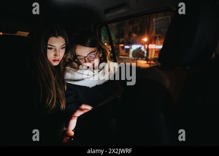 Deux jeunes femmes regardent un écran de téléphone intelligent tout en étant assis ensemble sur le siège arrière d'une voiture pendant une promenade de nuit dans la ville. Banque D'Images