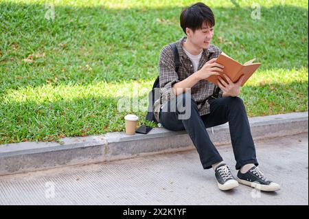 Un jeune étudiant asiatique positif et beau est assis dans la rue dans le parc du campus lisant un livre, profitant de ses loisirs en plein air. université l Banque D'Images