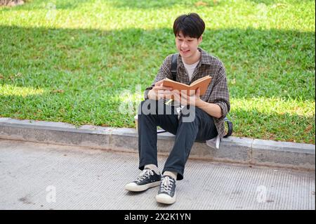 Un jeune étudiant asiatique positif et beau est assis dans la rue dans le parc du campus lisant un livre, profitant de ses loisirs en plein air. université l Banque D'Images