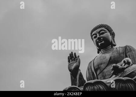Une photographie frappante en noir et blanc d'une grande statue de Bouddha, représentée avec une expression paisible et la main levée, posée contre un ciel nuageux. Banque D'Images