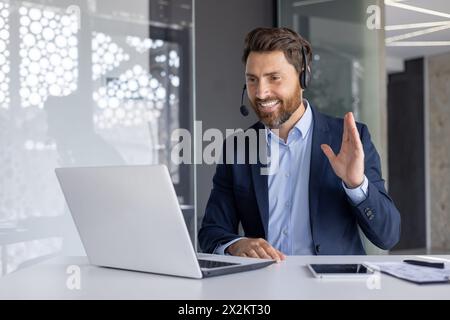 Un professionnel joyeux en costume participe à une conférence téléphonique en ligne à l'aide d'un casque, agitant la main, éventuellement en saluant ou en expliquant un point, dans un bureau moderne et bien éclairé. Banque D'Images