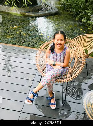 Portrait d'une fille asiatique assise dans un jardin avec fond d'étang, vue d'en haut, lumière du jour, image verticale. Banque D'Images