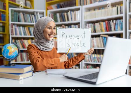 Une femme musulmane joyeuse en hijab démontre la grammaire anglaise, en utilisant «present simple» sur un tableau blanc dans une bibliothèque colorée. Banque D'Images