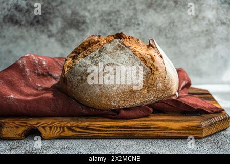 Le pain au levain de seigle fraîchement cuit repose sur une planche à découper en bois rustique, drapé d'un tissu Bordeaux sur un fond gris moucheté Banque D'Images